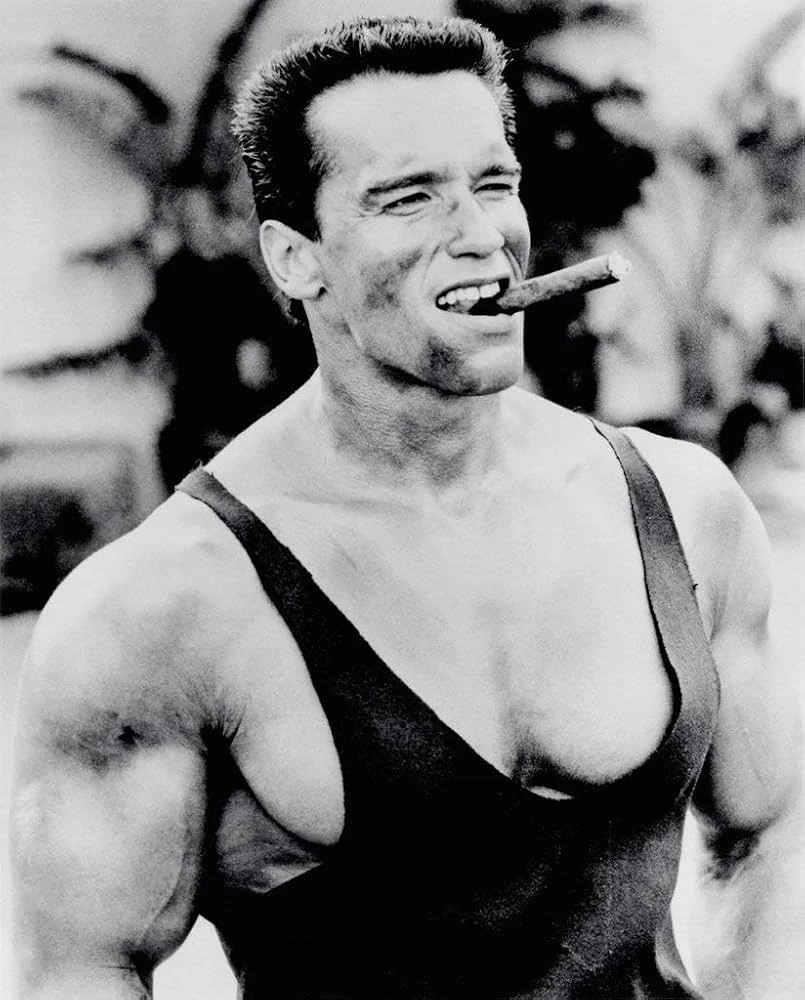 Arnold Schwarzenegger smiling with a cigar between his teeth, wearing a dark tank top, in a black-and-white photograph